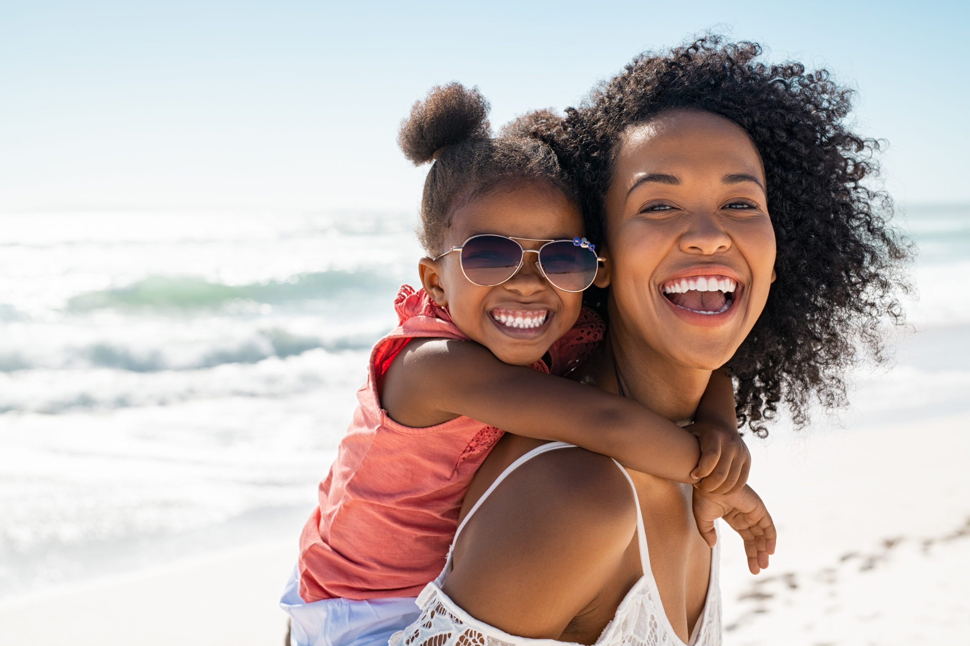 Happy young mother giving laughing daughter piggyback ride at beach Smiling black mother and beautiful daughter wearing sunglasses having fun on the beach. Portrait of happy african american woman giving a piggyback ride to her cute little girl wearing shades while looking at camera. Kid embracing her bigger sister during summer vacation with copy space.